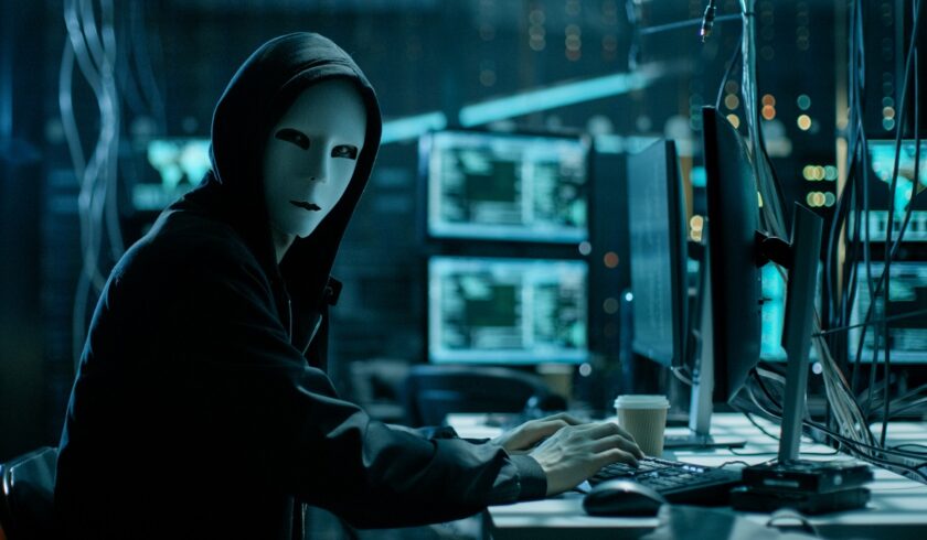 Hacker care are mască albă pe față, îmbrăcat într-o bluză cu glugă neagră, stă la un birou cu multe calculatoare în jur. Cea mai mare amenințare de securitate cibernetică a îngrijorat experții