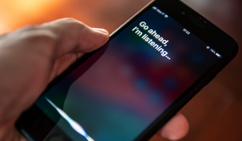 Un utilizator care are un iPhone în mână și vorbește cu Siri pe ecran. „O mică parte” din dispozitivele iPhone au înregistrat interacțiunile cu Siri