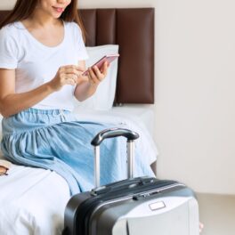 Femeie care stă pe pat, lângă bagaje, în camera de hotel. Experții au dezvăluit cum îți poți proteja lucrurile din camera de hotel
