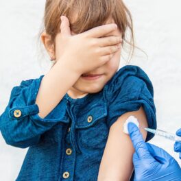 Fetiță care e vaccinată și care ține mână pe ochi, într-o rochie albastră, pe fundal alb. Pojarul e un patogen antic, care a fost declarat pericol iminent