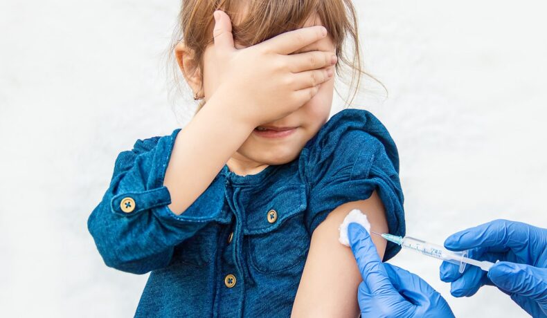 Fetiță care e vaccinată și care ține mână pe ochi, într-o rochie albastră, pe fundal alb. Pojarul e un patogen antic, care a fost declarat pericol iminent
