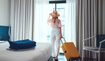 Femeia care e într-o cameră de hotel, cu o valiză portocalie, se uită pe geam, lângă un pat. Stewardesele au dezvăluit trucuri pentru a te proteja în camera de hotel
