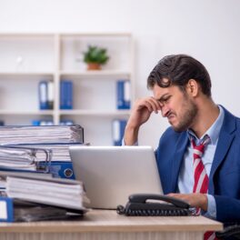 Bărbat îmbrăcat în costum albastru, care stă la un birou plin de documente, pentru a ilustra cele mai „nefericite joburi” din lume