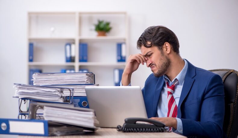 Bărbat îmbrăcat în costum albastru, care stă la un birou plin de documente, pentru a ilustra cele mai „nefericite joburi” din lume