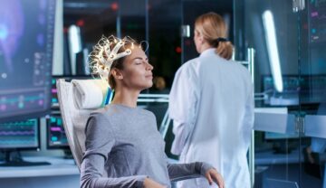 Femeie care stă în scaun, cu electrozi pe cap, cu o doctoriță lângă, pe fundal un laborator albastru, pentru a ilustra inteligența artificială care poate „citi gânduri”