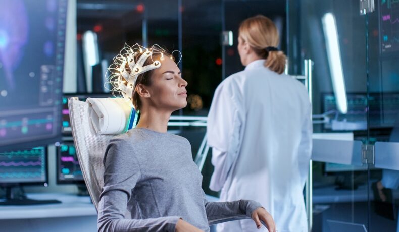 Femeie care stă în scaun, cu electrozi pe cap, cu o doctoriță lângă, pe fundal un laborator albastru, pentru a ilustra inteligența artificială care poate „citi gânduri”