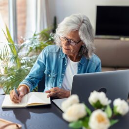 Femeie în vârstă, cu părul alb și cămașă albastră, care stă la birou, în fața unui laptop, care scrie într-un jurnal, pentru a ilustra o „mică bunicuță” care riscă 10 ani de închisoare