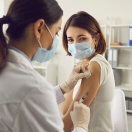 Femeie care e vaccinată de o doctoriță în mâna stângă, ambele purtând mască, pentru a ilustra ce e „boala x”