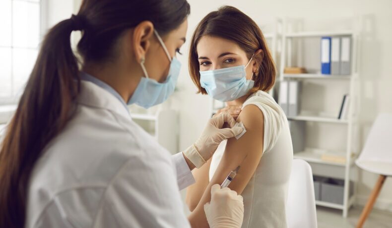 Femeie care e vaccinată de o doctoriță în mâna stângă, ambele purtând mască, pentru a ilustra ce e „boala x”
