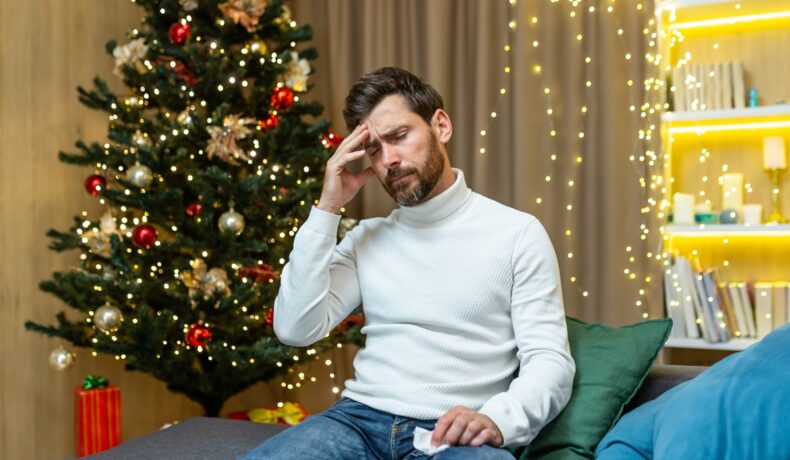 Bărbat care are „sindromul bradului de Crăciun”, îmbrăcat în pulover alb, pe o canapea, cu pomul de Crăciun decorat pe fundal
